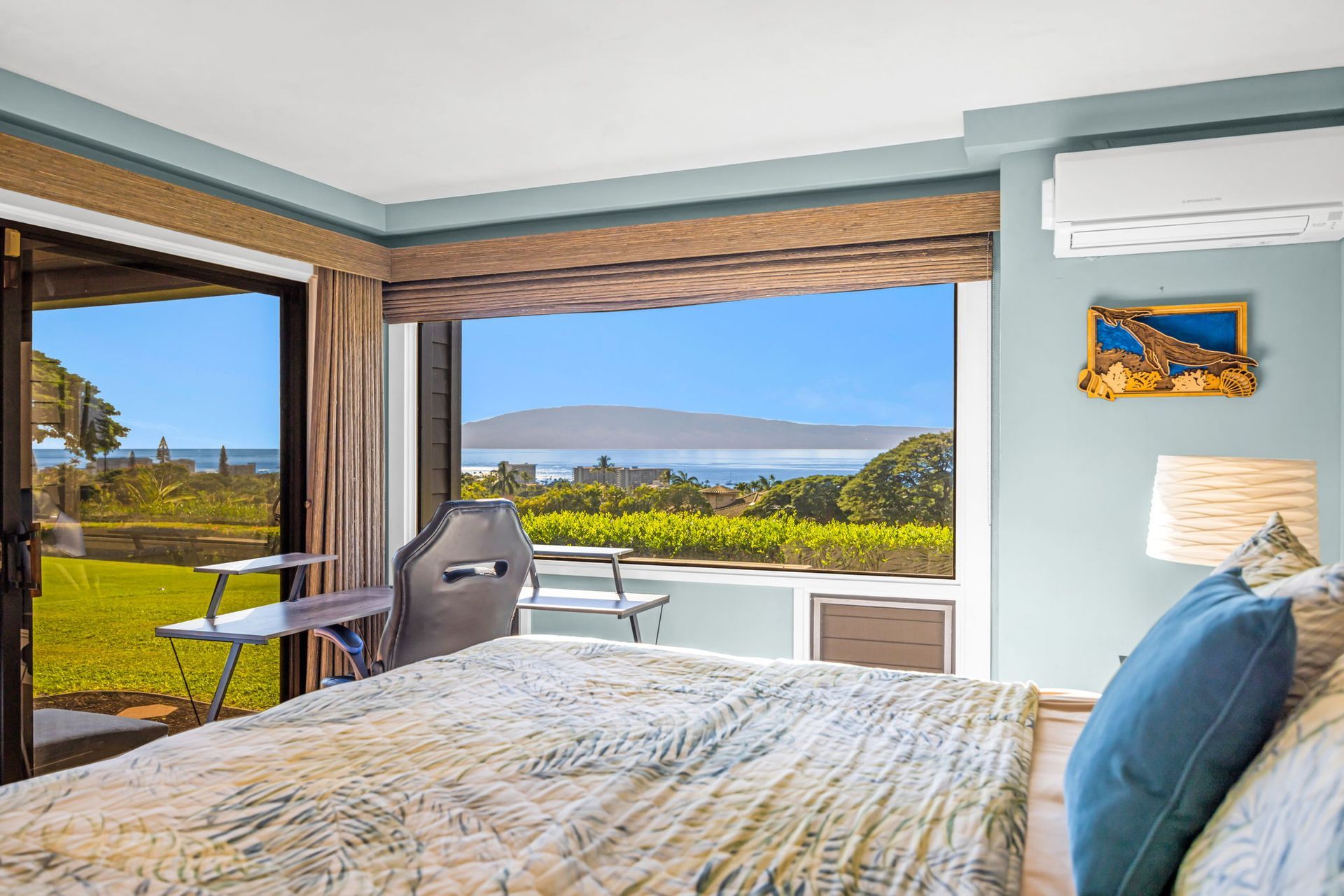 A sunlit bedroom overlooking a coastal view, featuring a bed with a patterned quilt, a desk, and wall art.