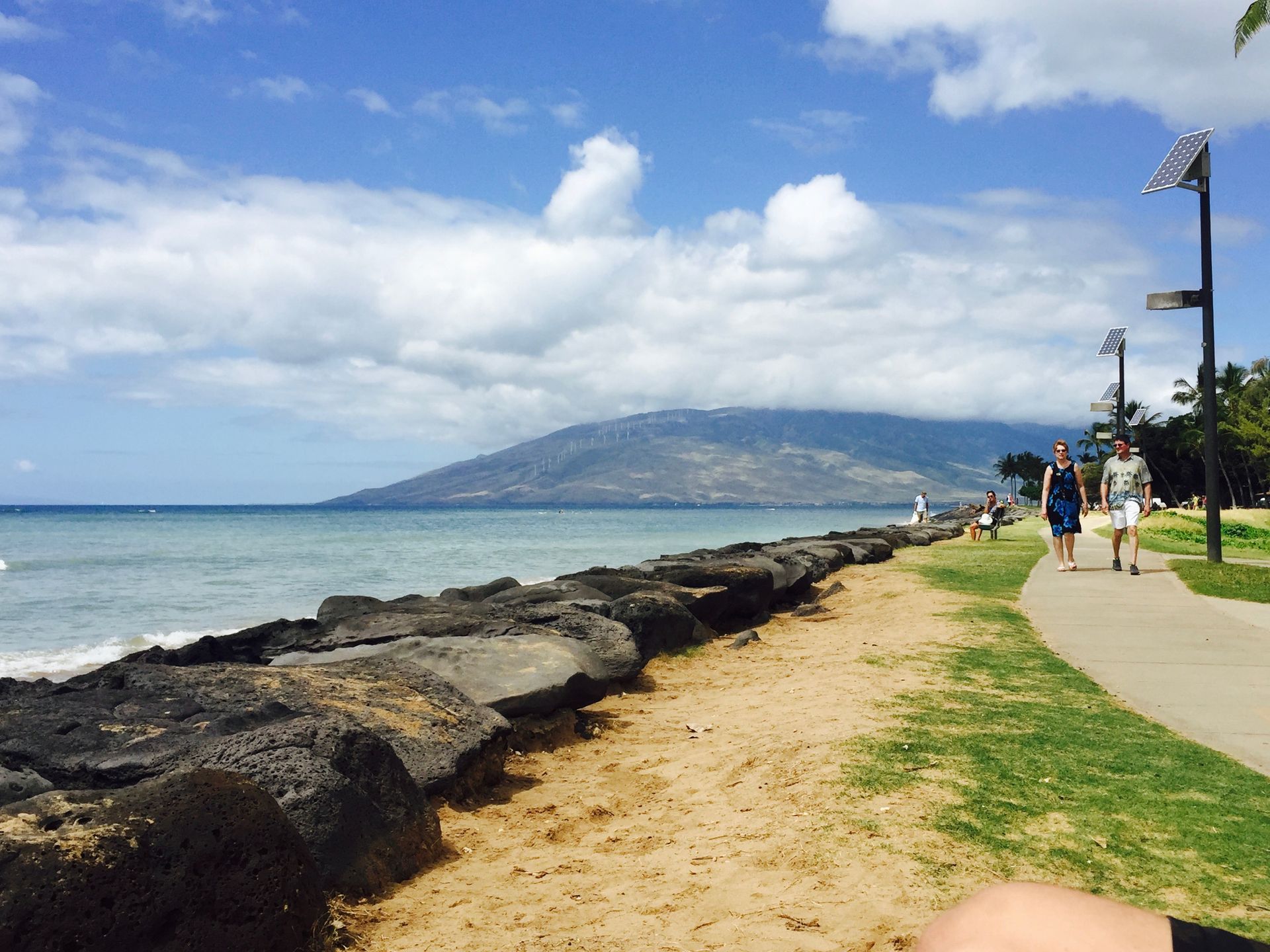 A paved path runs beside a black volcanic rock seawall and ocean, with a mountain in the distance under a cloudy sky.