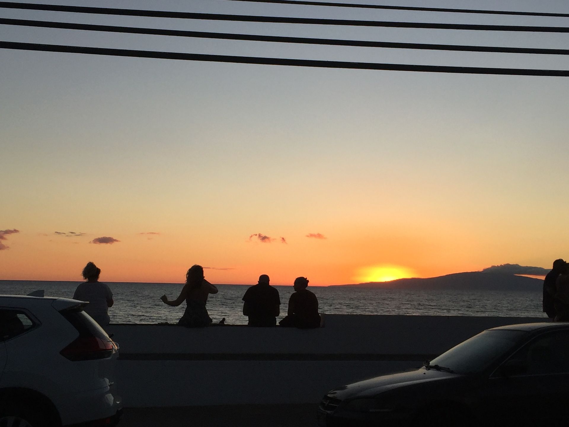 Four people sit along a seawall at sunset, silhouetted against an orange sky and the ocean, with cars in the foreground.