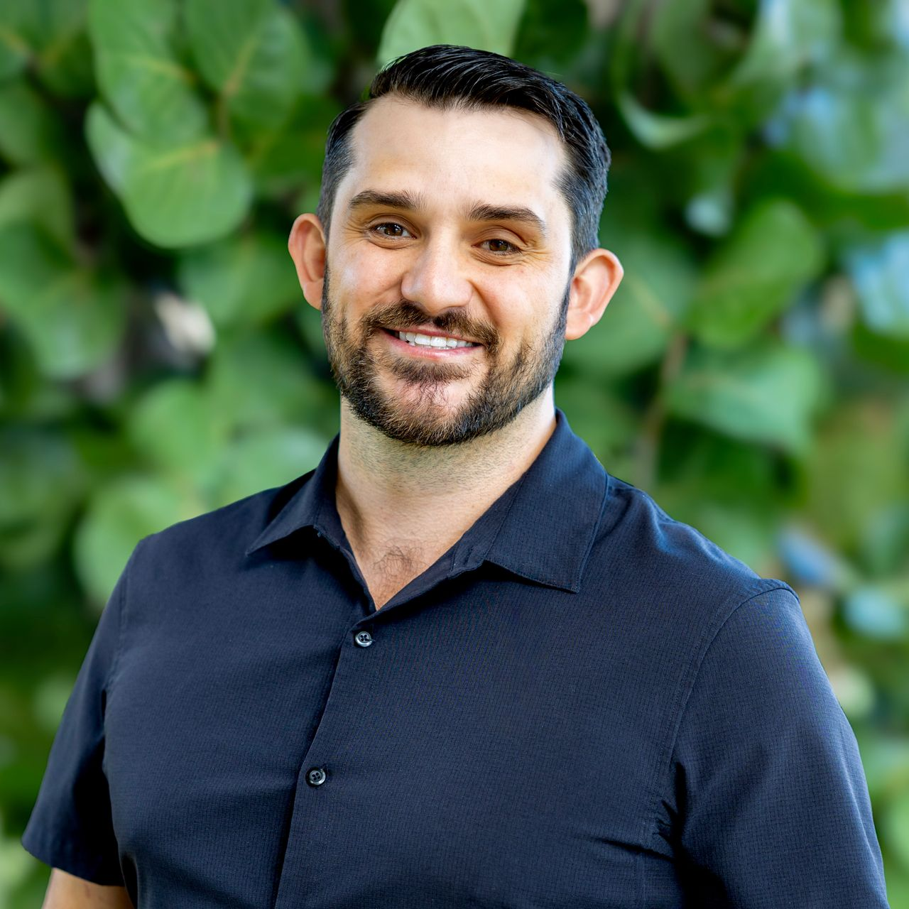 A person with a beard and dark hair smiles, wearing a navy short-sleeved collared shirt in front of a blurry green backdrop.