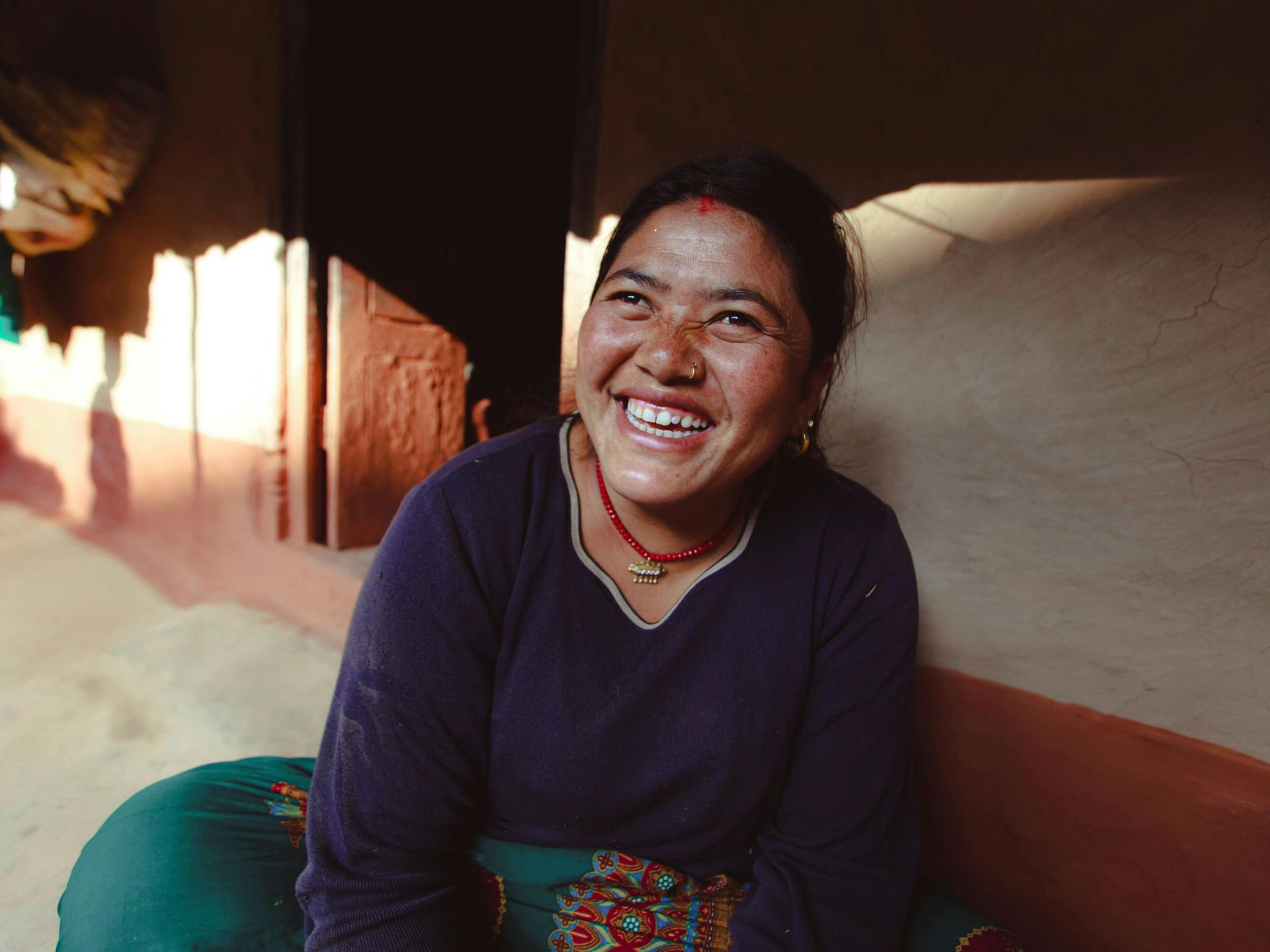 A smiling person in a dark blue sweater and red beaded necklace, sitting against a textured, light-colored wall.