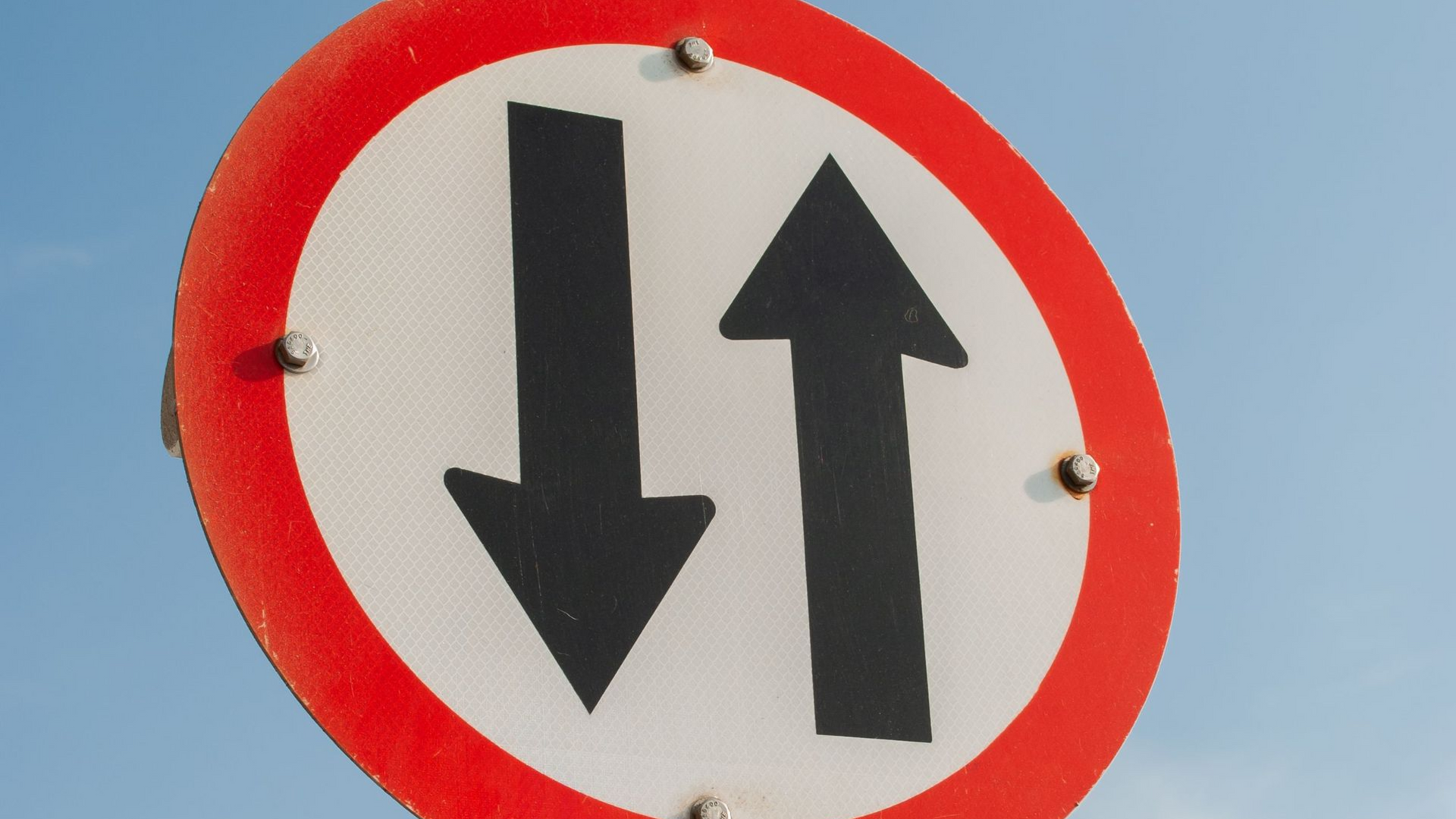 Round traffic sign with a red border and black up and down arrows on a blue sky background