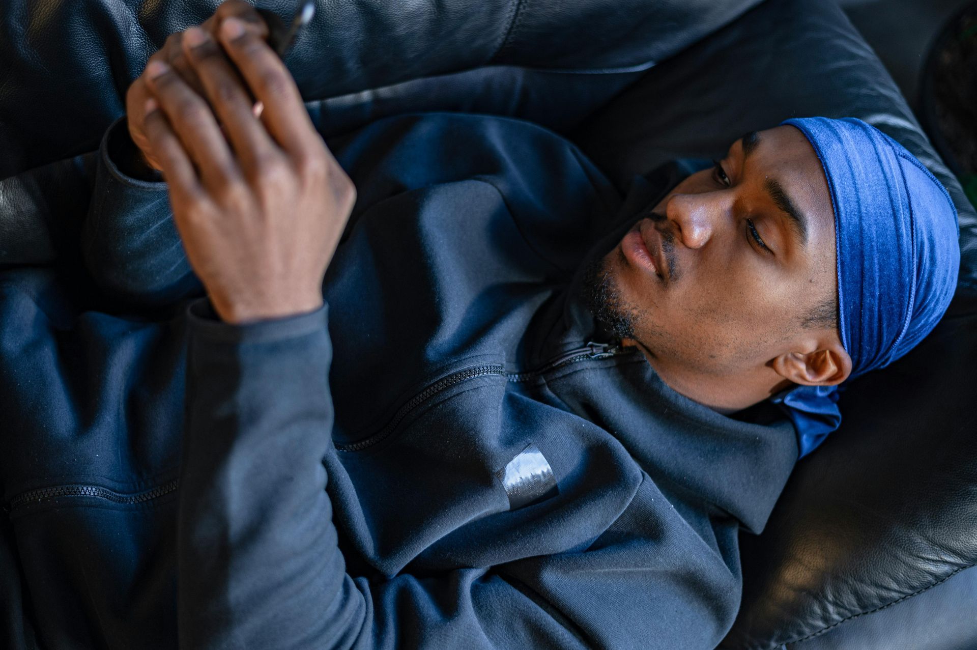 A person wearing a blue durag and dark zip-up jacket reclines while looking down at a smartphone.
