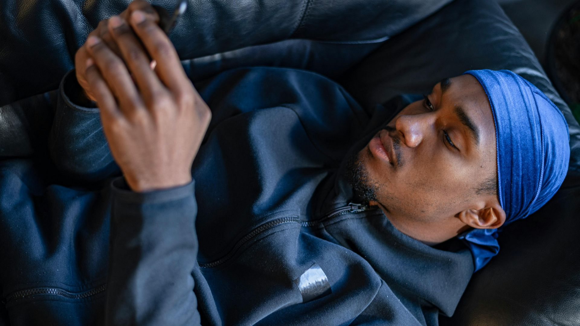A person wearing a blue durag and dark hoodie reclines while focused on their smartphone.