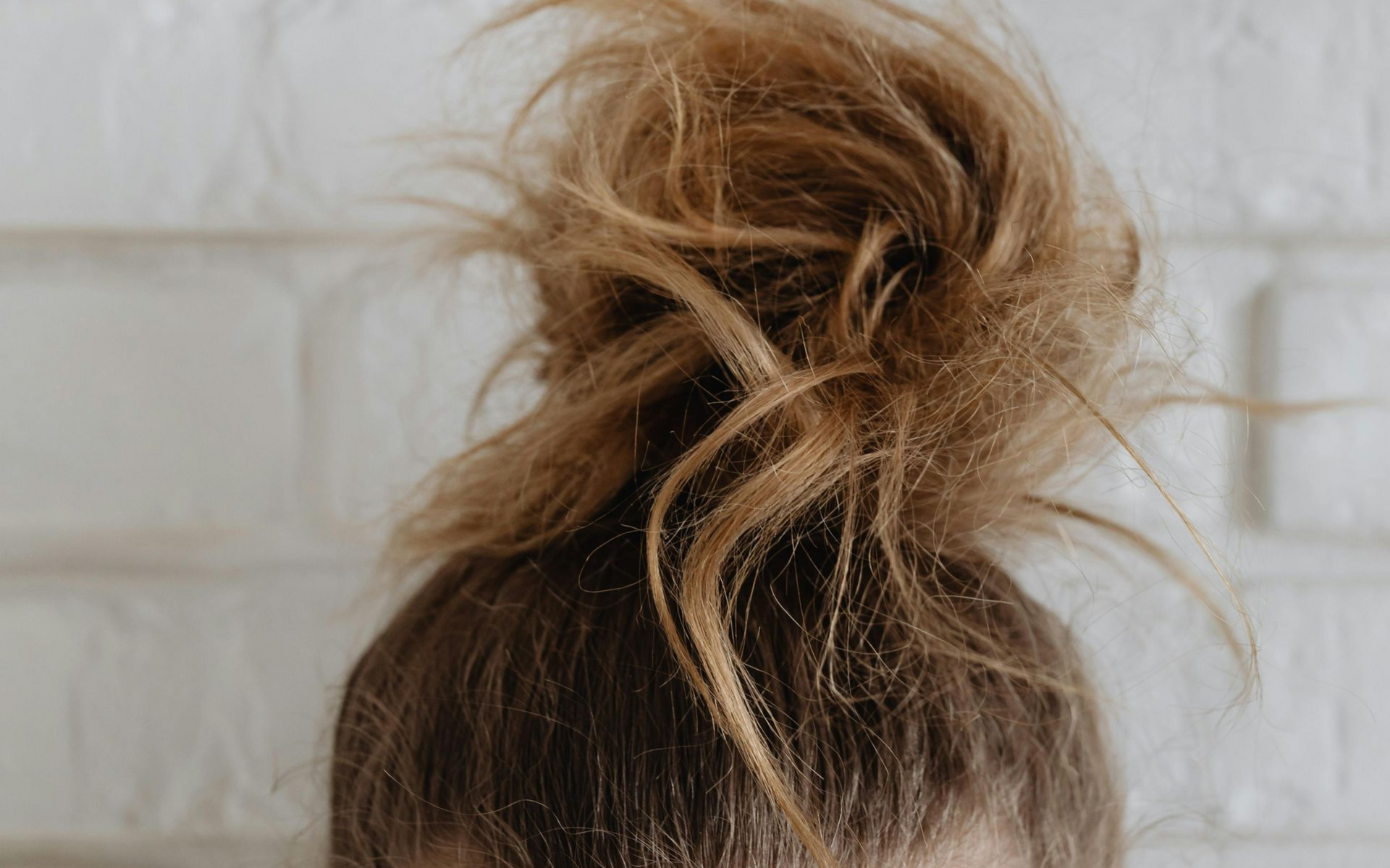 A messy, light brown hair bun styled high on top of a head against a white brick wall background.