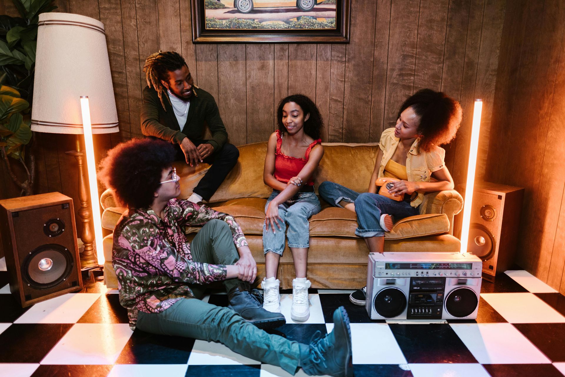 Four people relax in a cozy room with a checkered floor, warm lights, and a boombox on the floor.