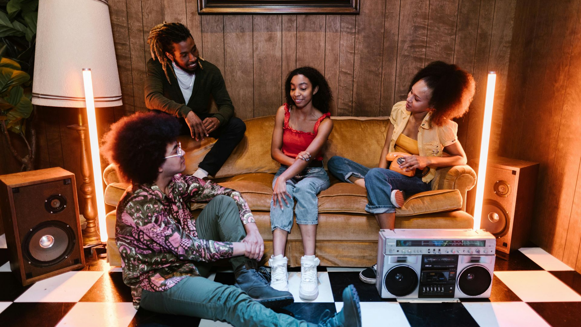Four people relax in a retro-style room with a gold sofa, wood-paneled walls, checkerboard floor, and a vintage boombox.