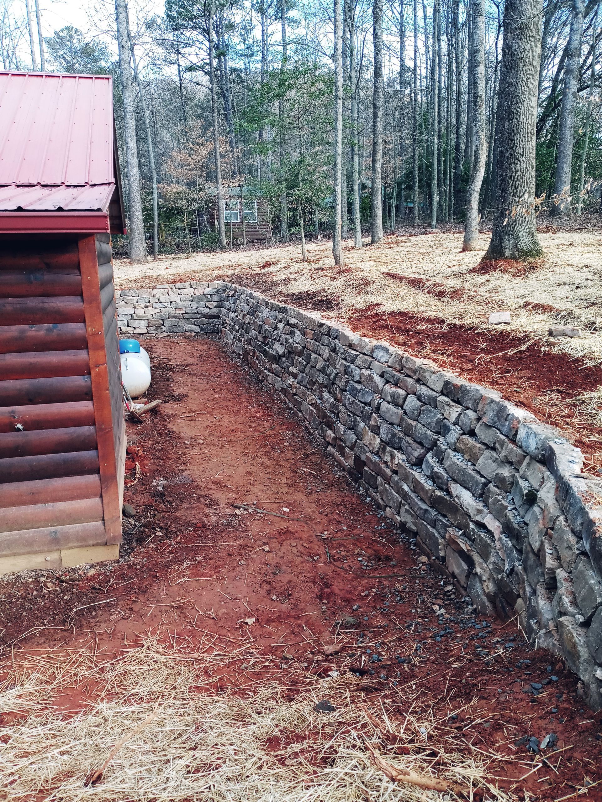 A log cabin is sitting next to a stone wall in the middle of a forest.