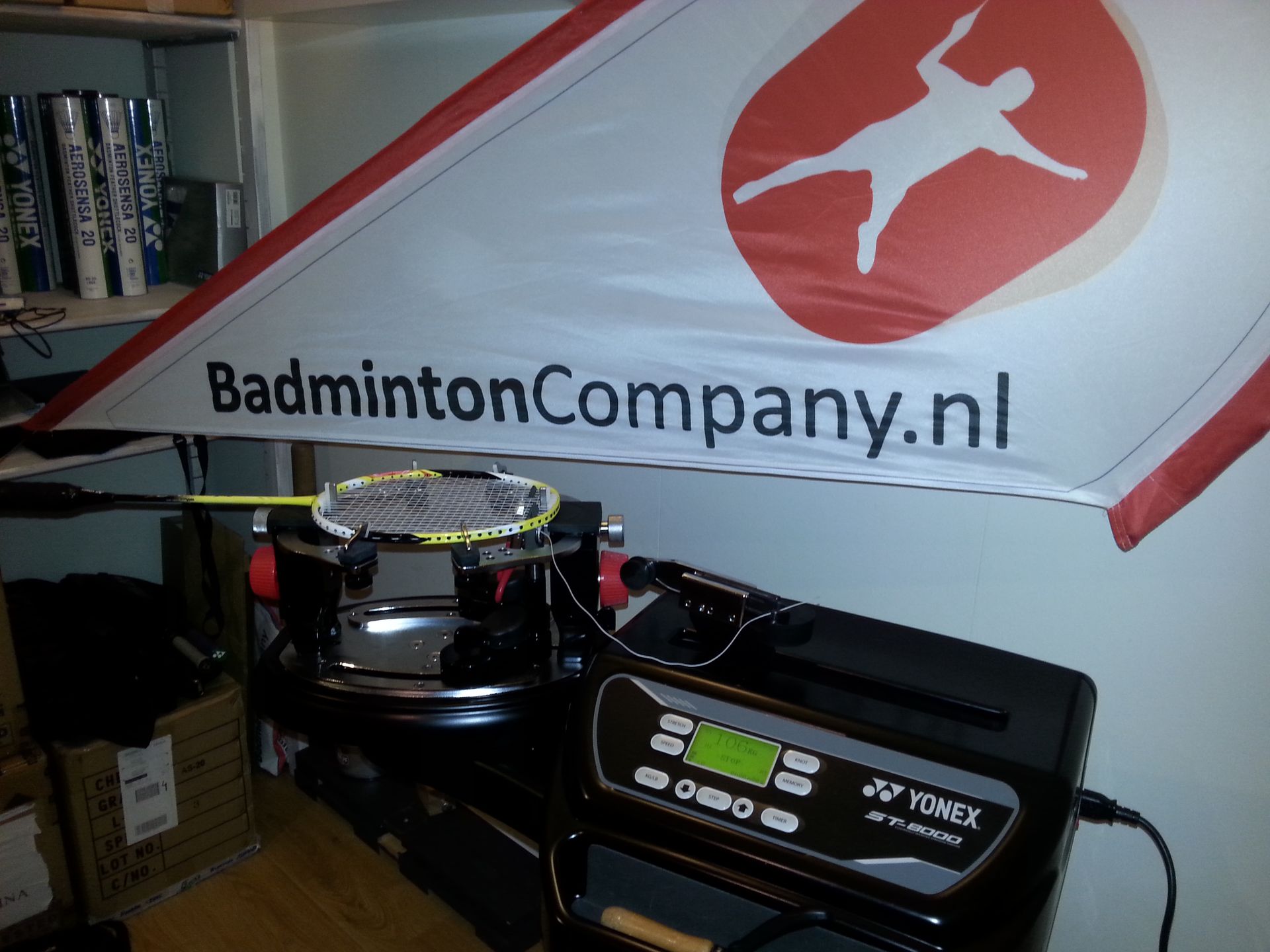 Een banner van BadmintonCompany.nl hangt boven een badmintonmachine en -uitrusting in een kamer.