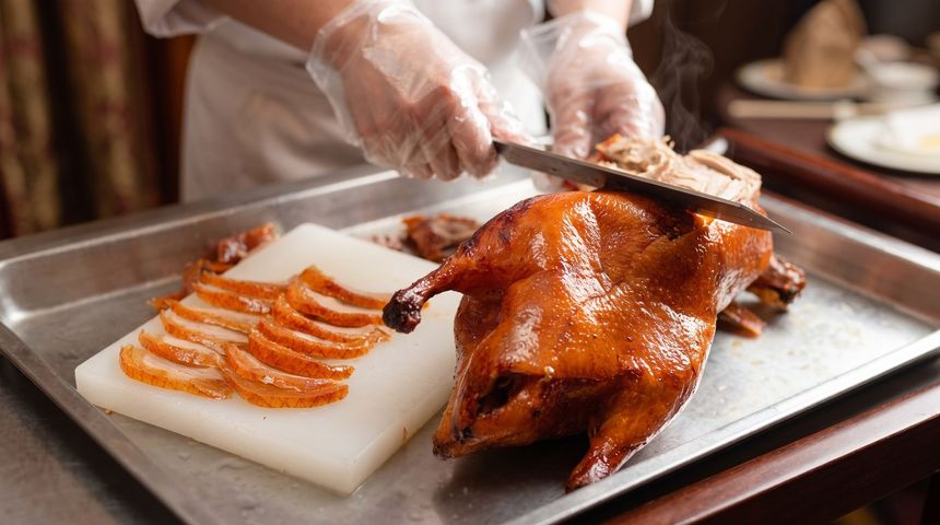 Chef carving a roasted duck on a tray beside neatly sliced pieces on a white cutting board.