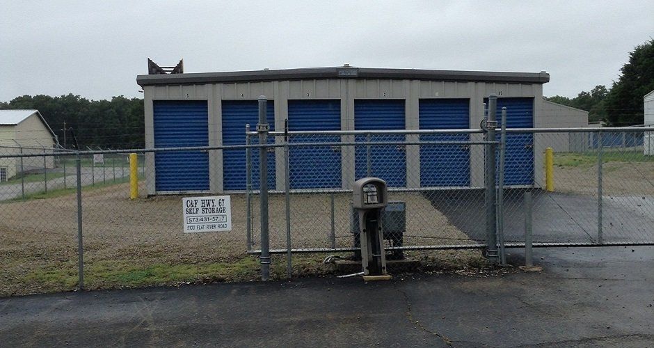 Small storage building with blue roll-up doors behind a chain-link fence on a gray day.