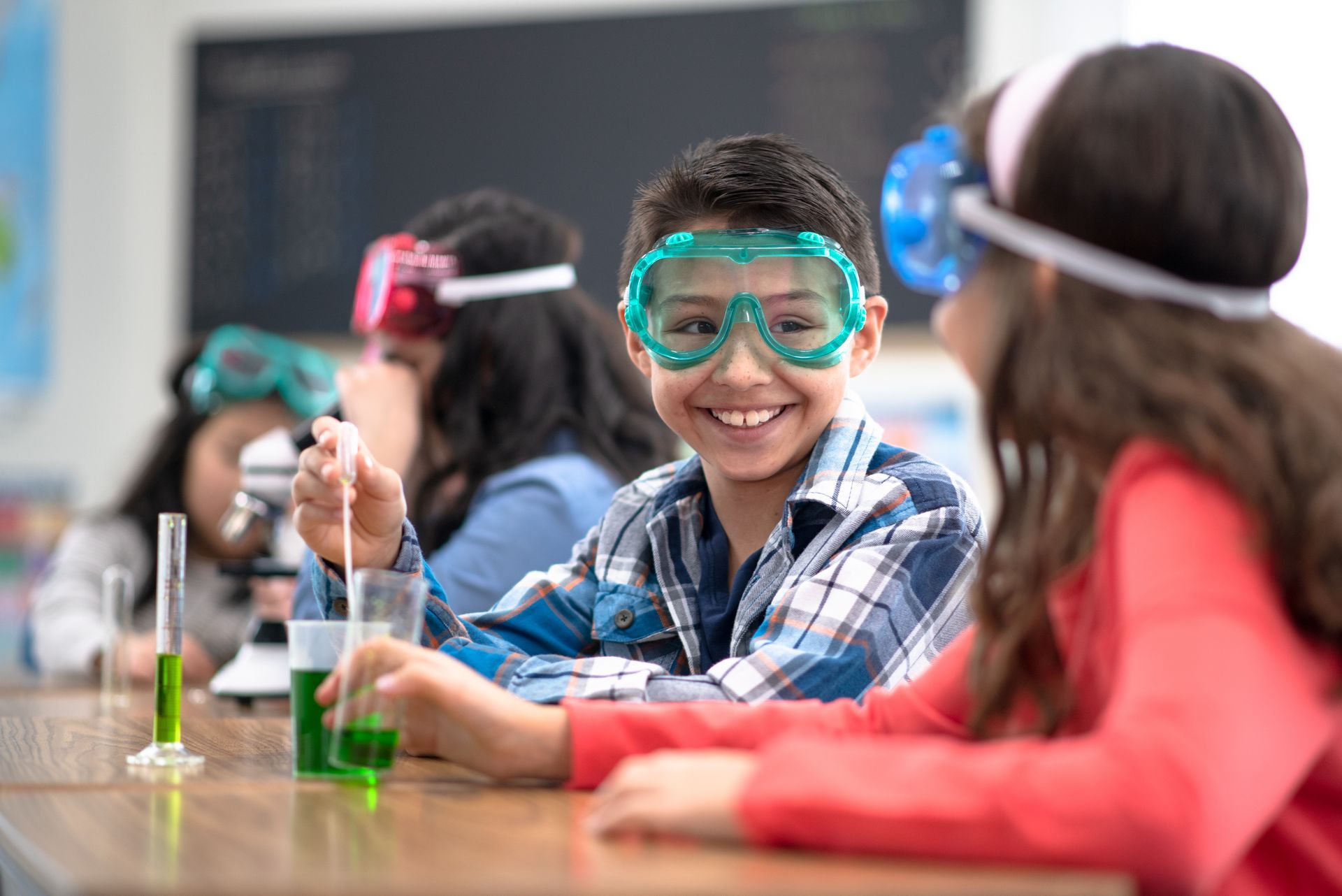 A boy and a girl wearing goggles are sitting at a table in a classroom.