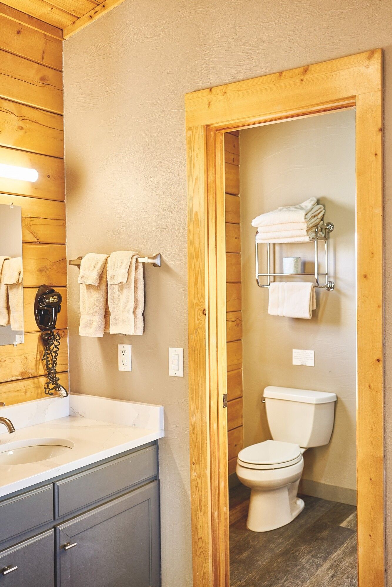 Rustic bathroom with wood paneling, sink, mirror, towels, and a toilet in a separate alcove.
