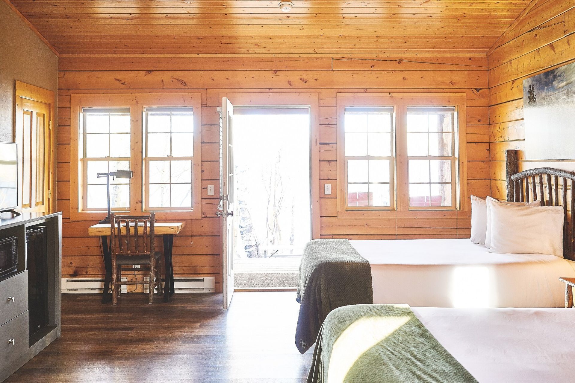 Cozy wooden cabin bedroom with two beds, desk by windows, and an open door letting in bright light
