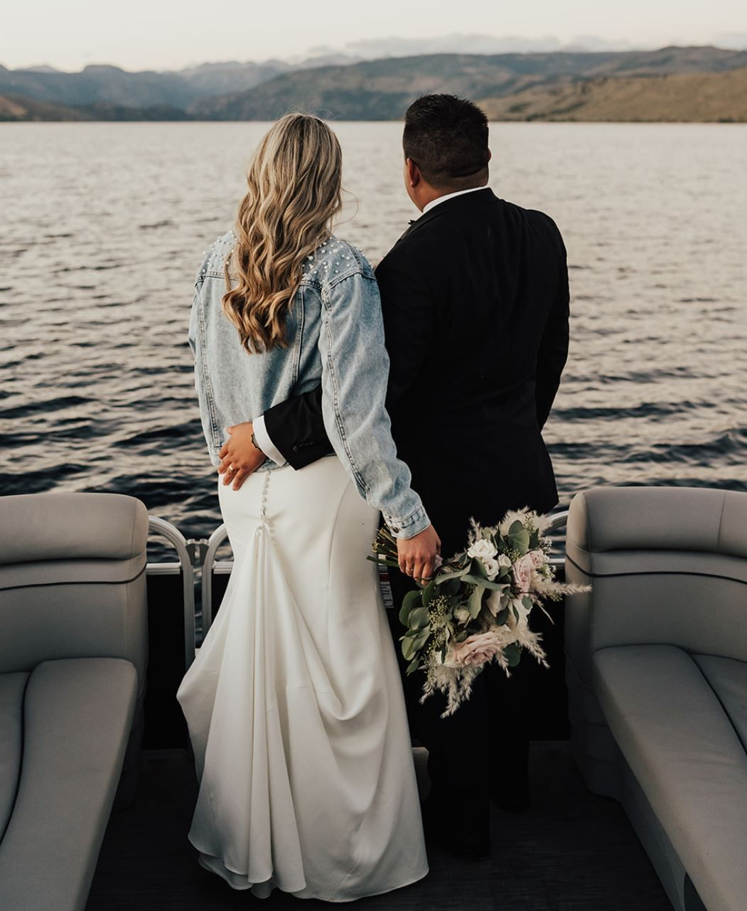 Couple standing on a boat, holding hands and facing a lake, with mountains in the distance.