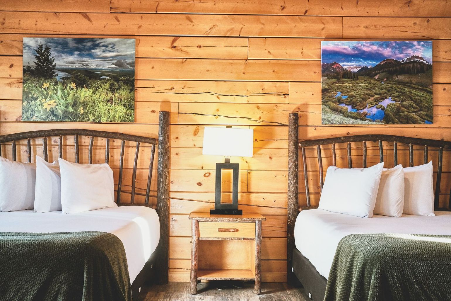 Rustic cabin bedroom with two beds, wooden walls, nightstand lamp, and framed landscape art
