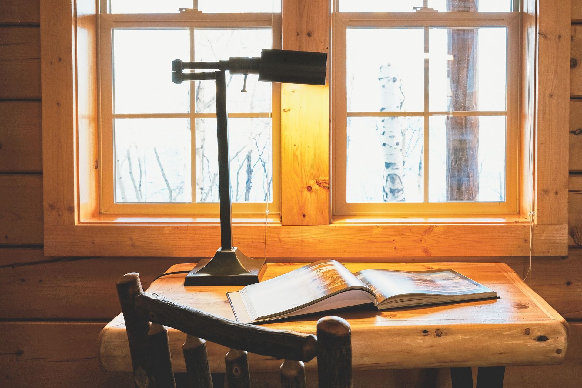 A sunlit desk by a window with a black lamp, open book, and chair in a cozy room.