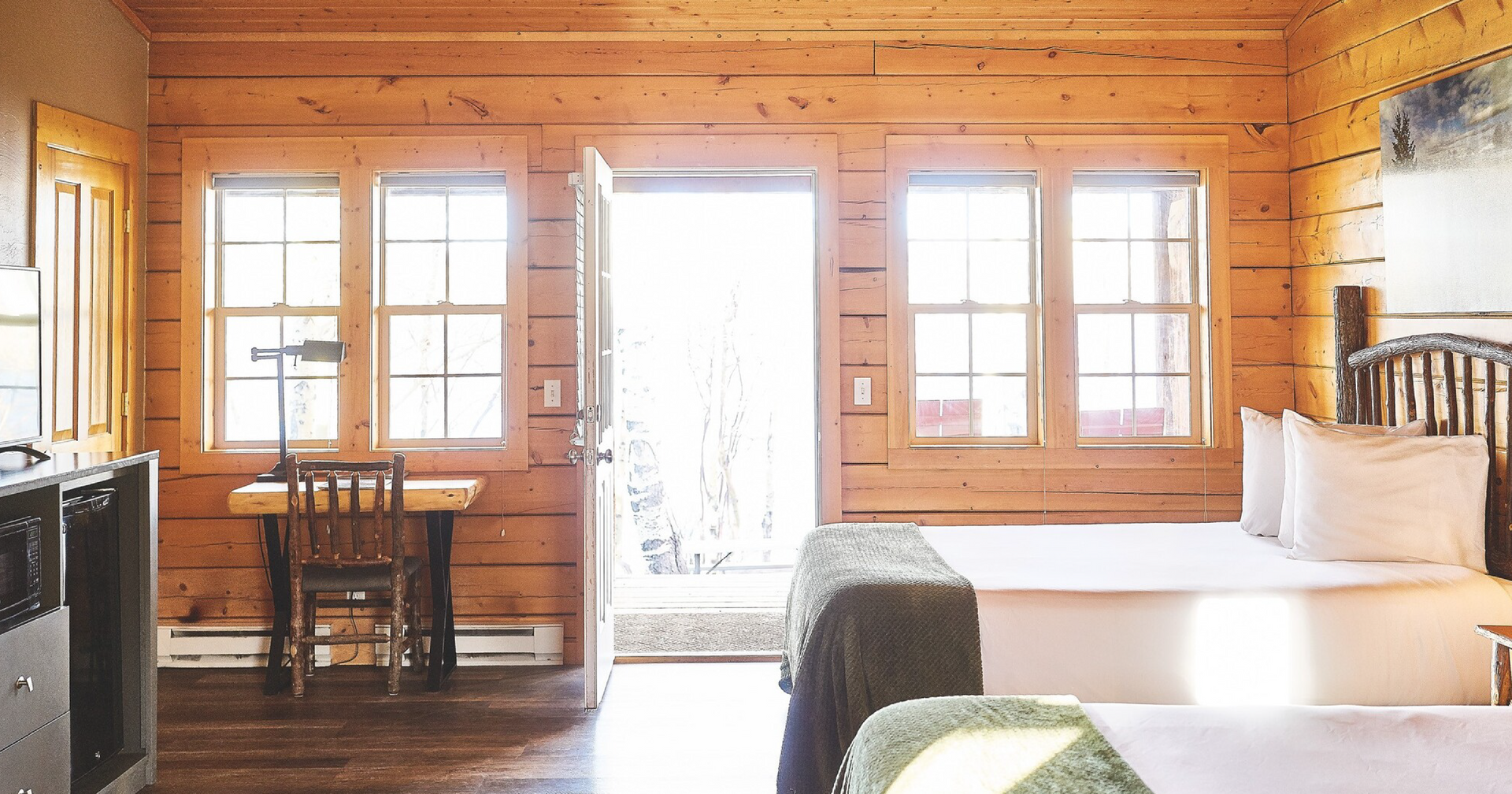 Wood-paneled cabin bedroom with twin beds, windows, a small table, and a bright open door