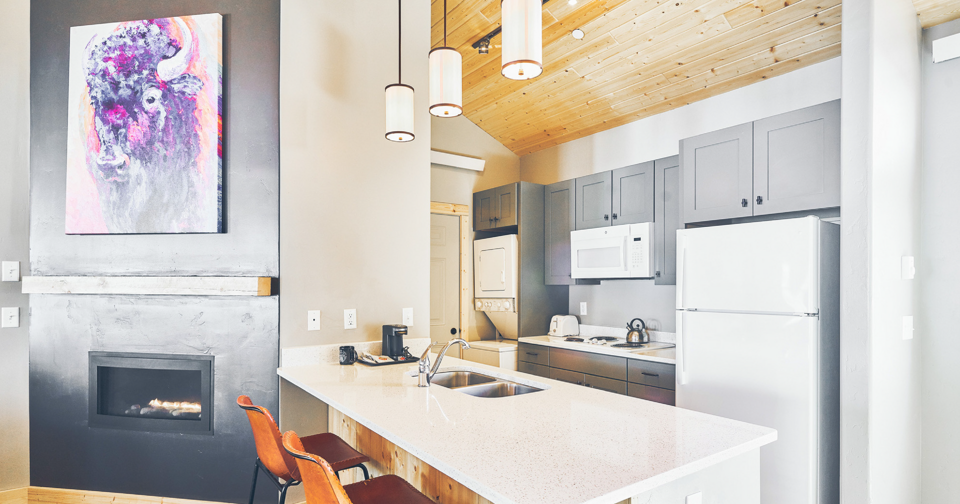 Modern kitchen with white island, gray cabinets, wood ceiling, pendant lights, and fireplace art.