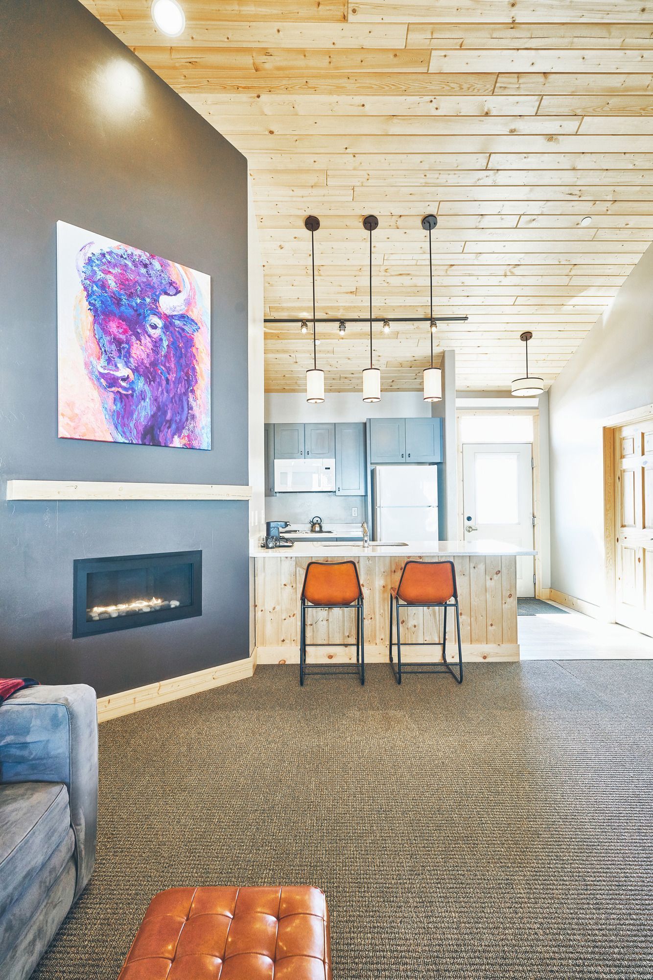Modern living room with fireplace, abstract art, kitchen island, and two orange stools beneath a wood ceiling