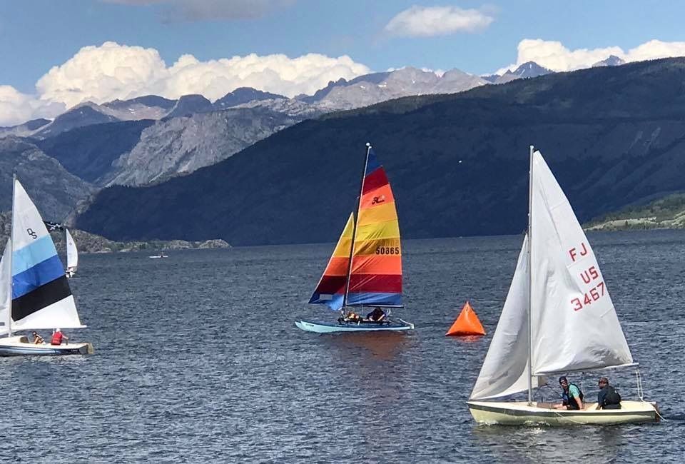 Three sailboats on a lake with mountains in the background, including a rainbow sailboat in the center.