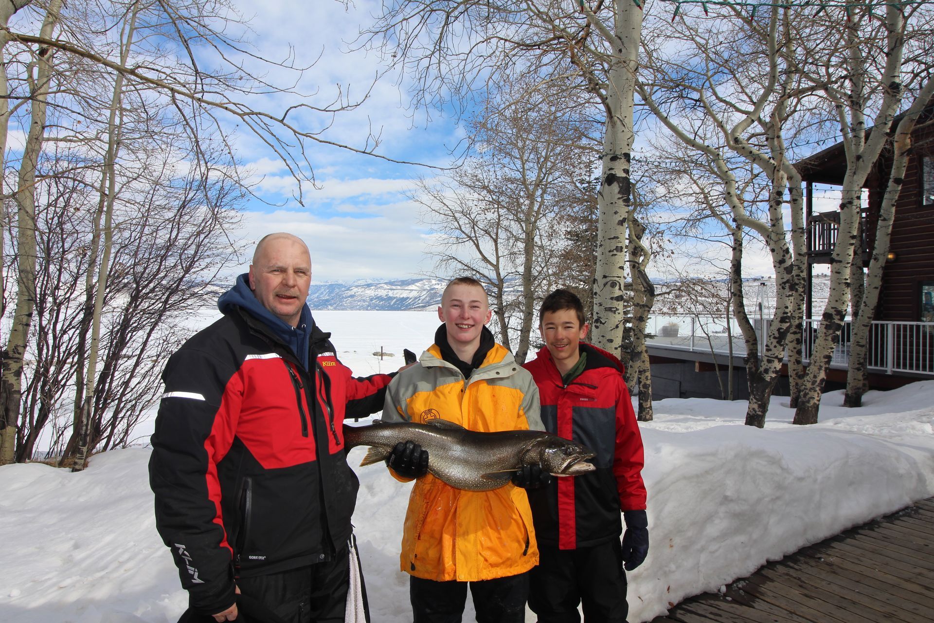 Three people in winter jackets holding a fish beside a snowy lakeside path