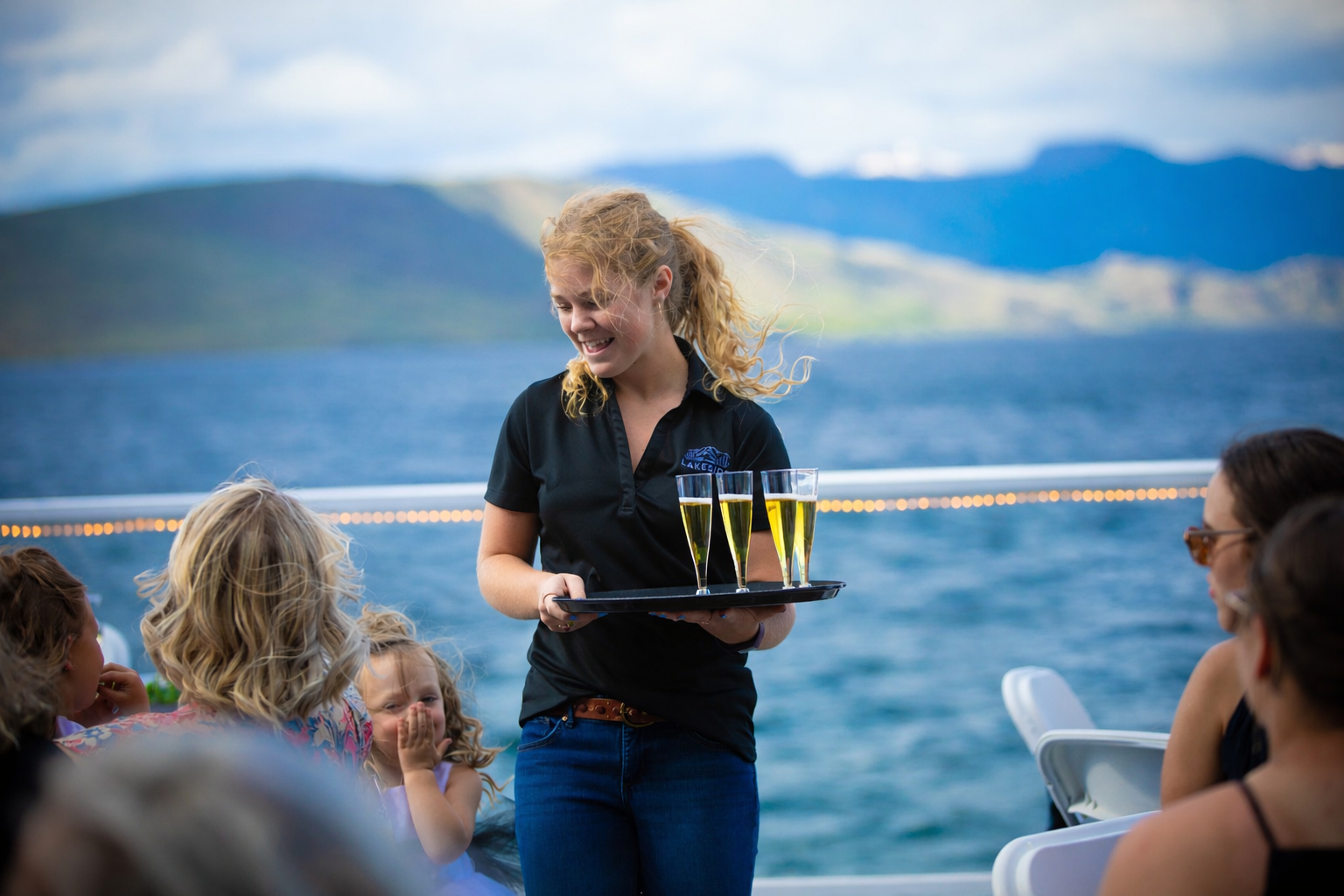 Server carrying drinks on a boat deck with lake and mountains in the background