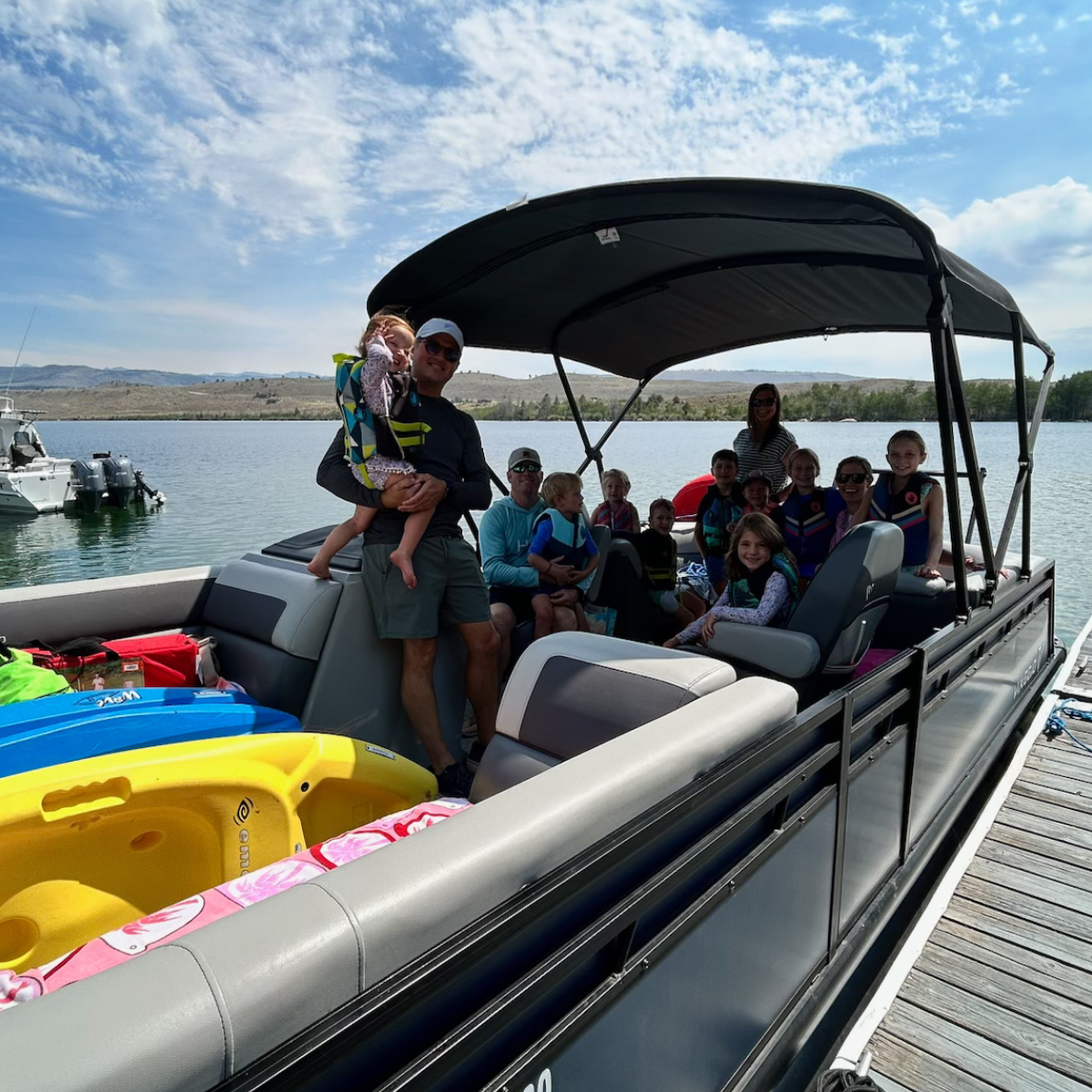Family on a pontoon boat with colorful floaties, cruising on a lake under a canopy
