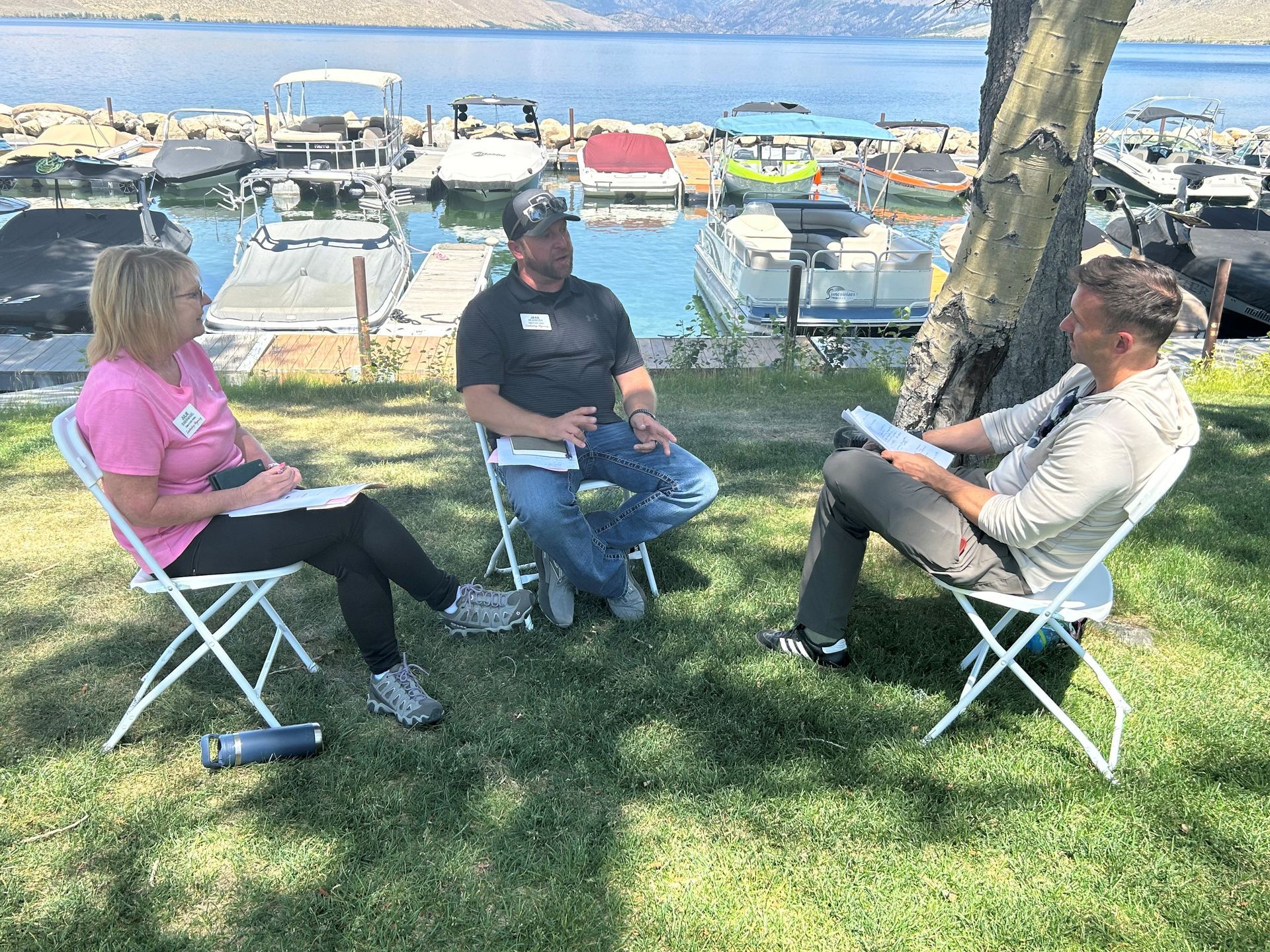 Three people sit on folding chairs in a grassy park by a marina, talking near trees and boats.