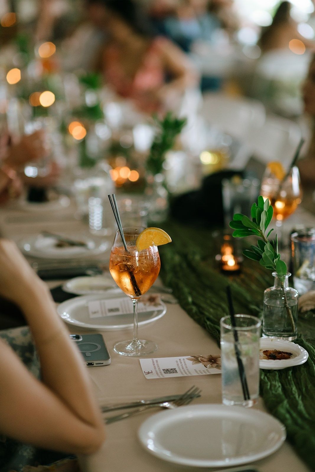 Elegant candlelit dining table with white plates, orange cocktails, and greenery centerpiece