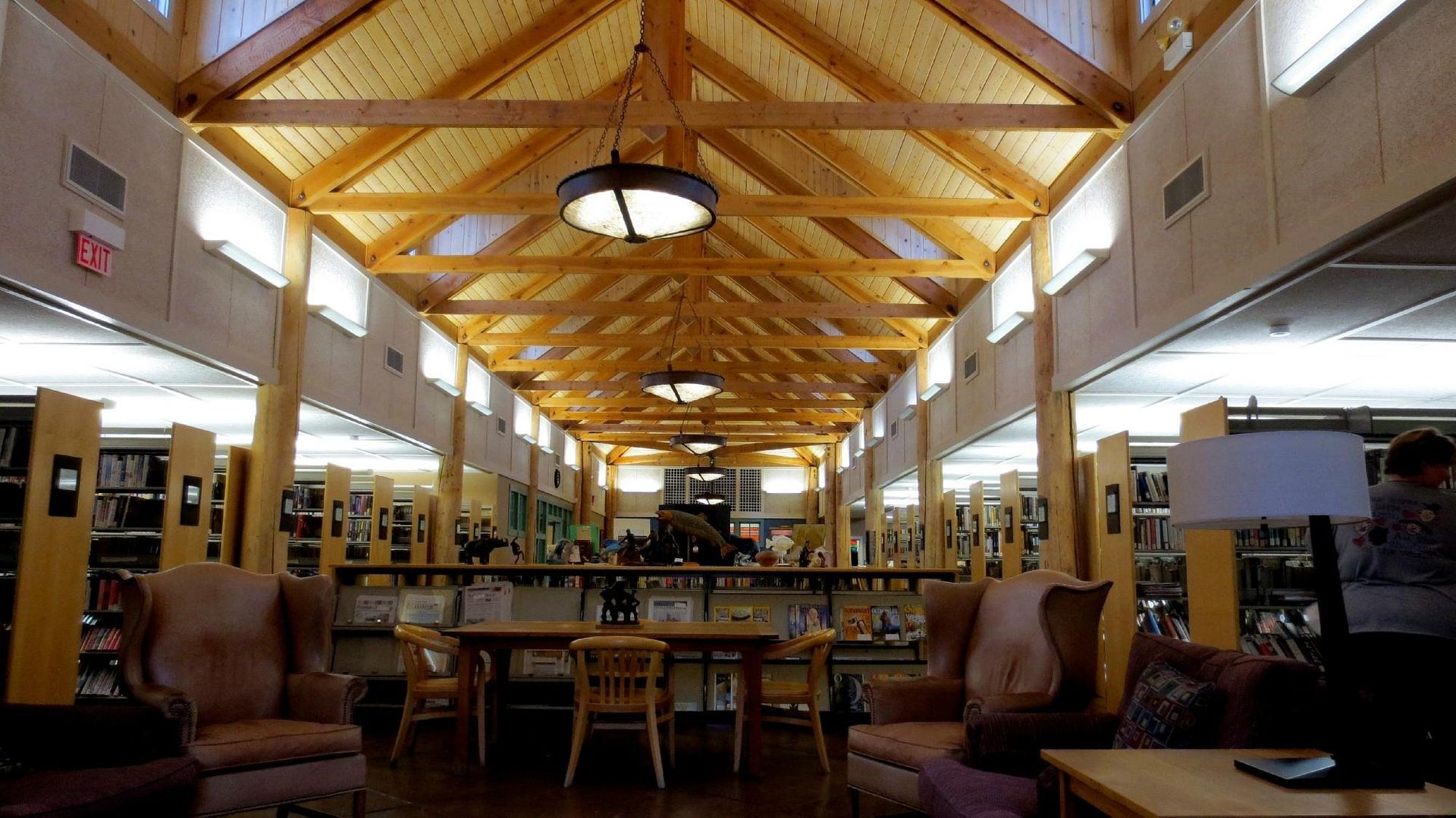 Spacious library reading room with wooden beams, hanging lights, tables, chairs, and bookshelves