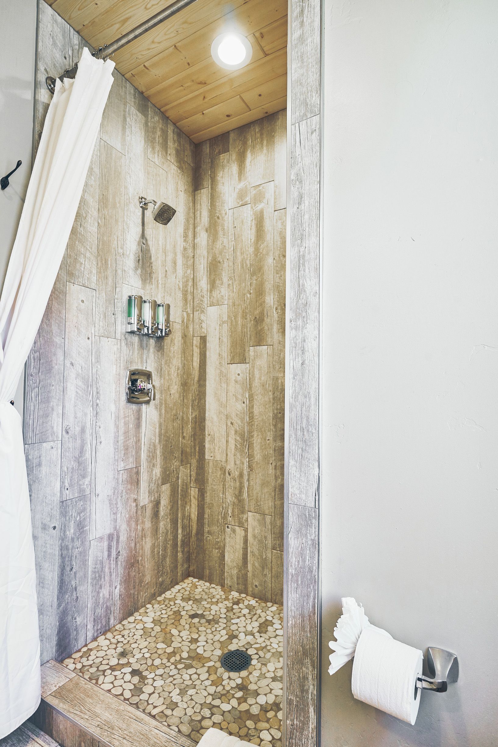 Small tiled shower stall with wood-look walls, overhead light, and a white towel on the side