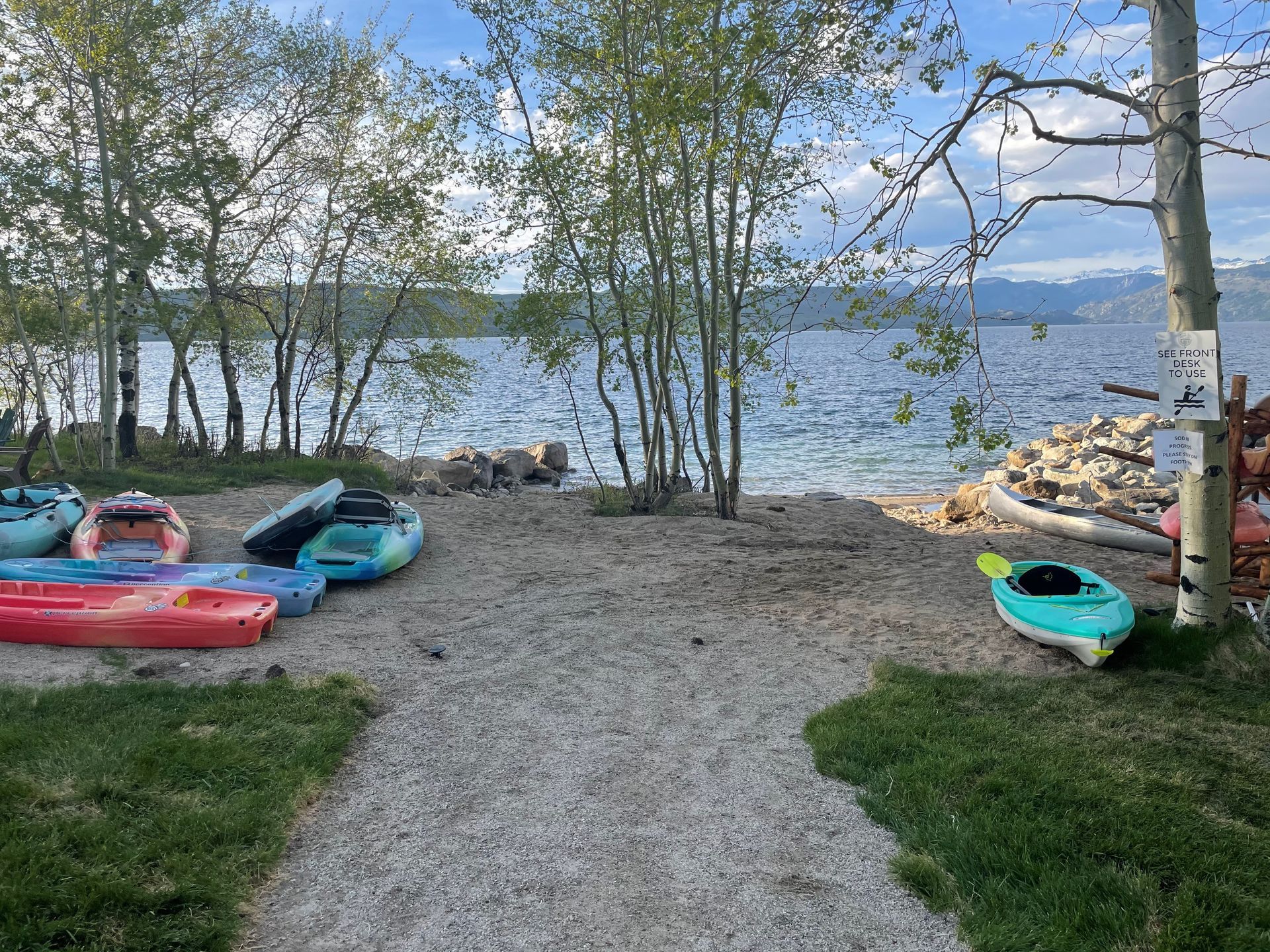 Kayaks on a gravel path by a lakeshore, with trees and calm water under a cloudy sky