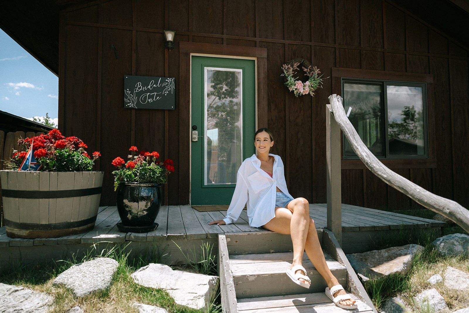 Person sitting on porch steps beside potted flowers outside a dark wooden cabin