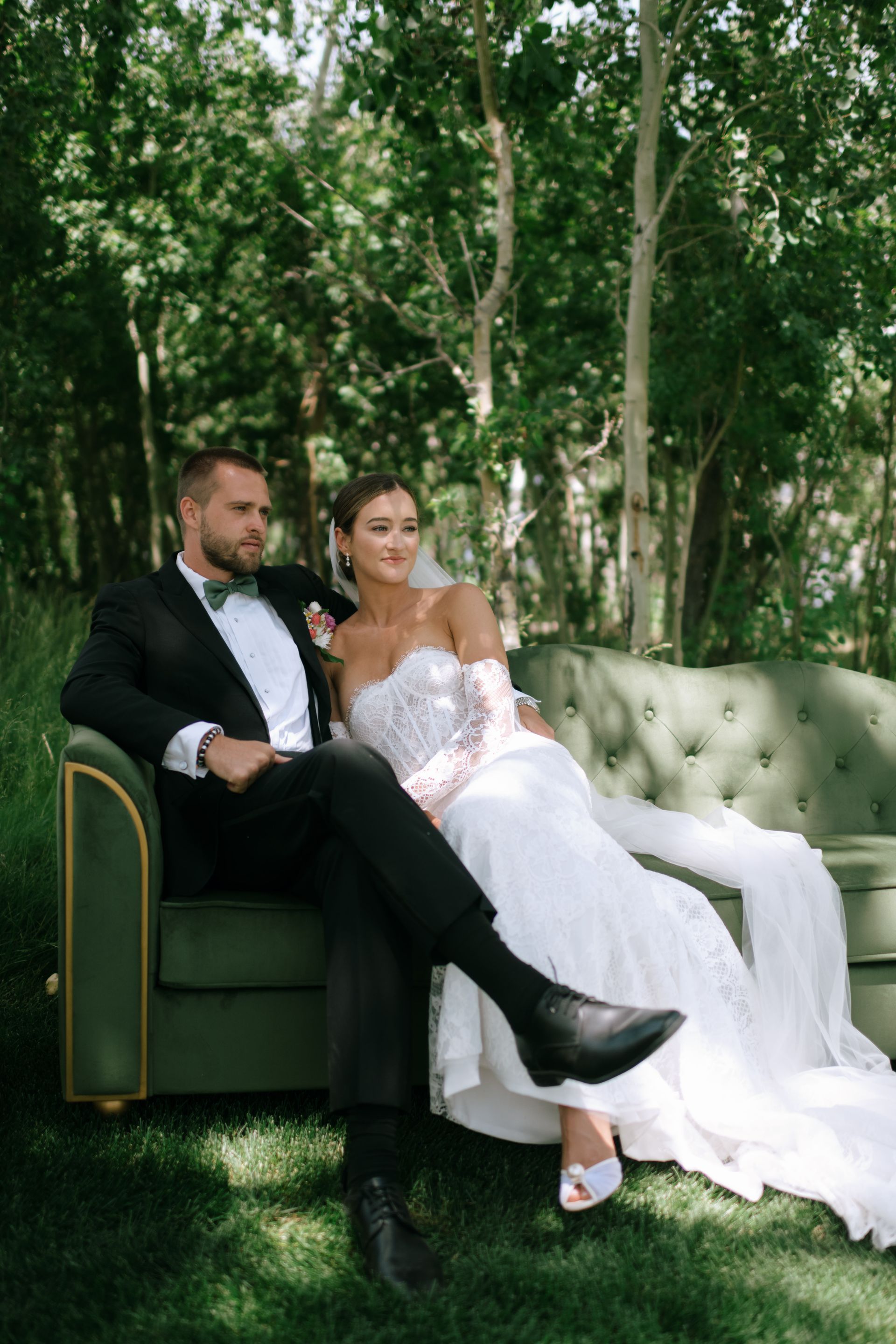 Couple in formalwear seated on a green velvet sofa in a wooded outdoor setting