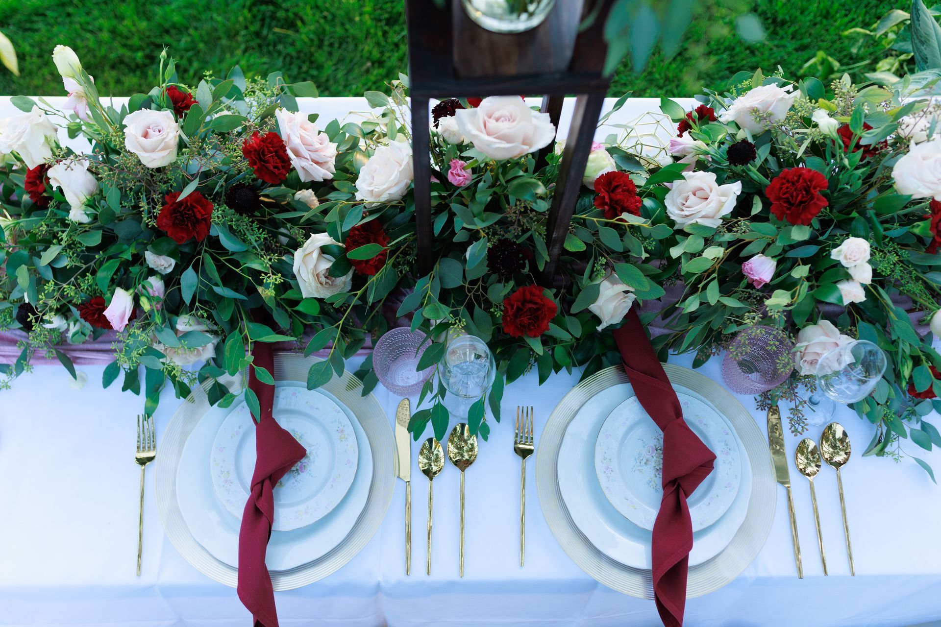 Elegant floral table arrangement with white and red roses, greenery, and burgundy ribbons on a white tablecloth