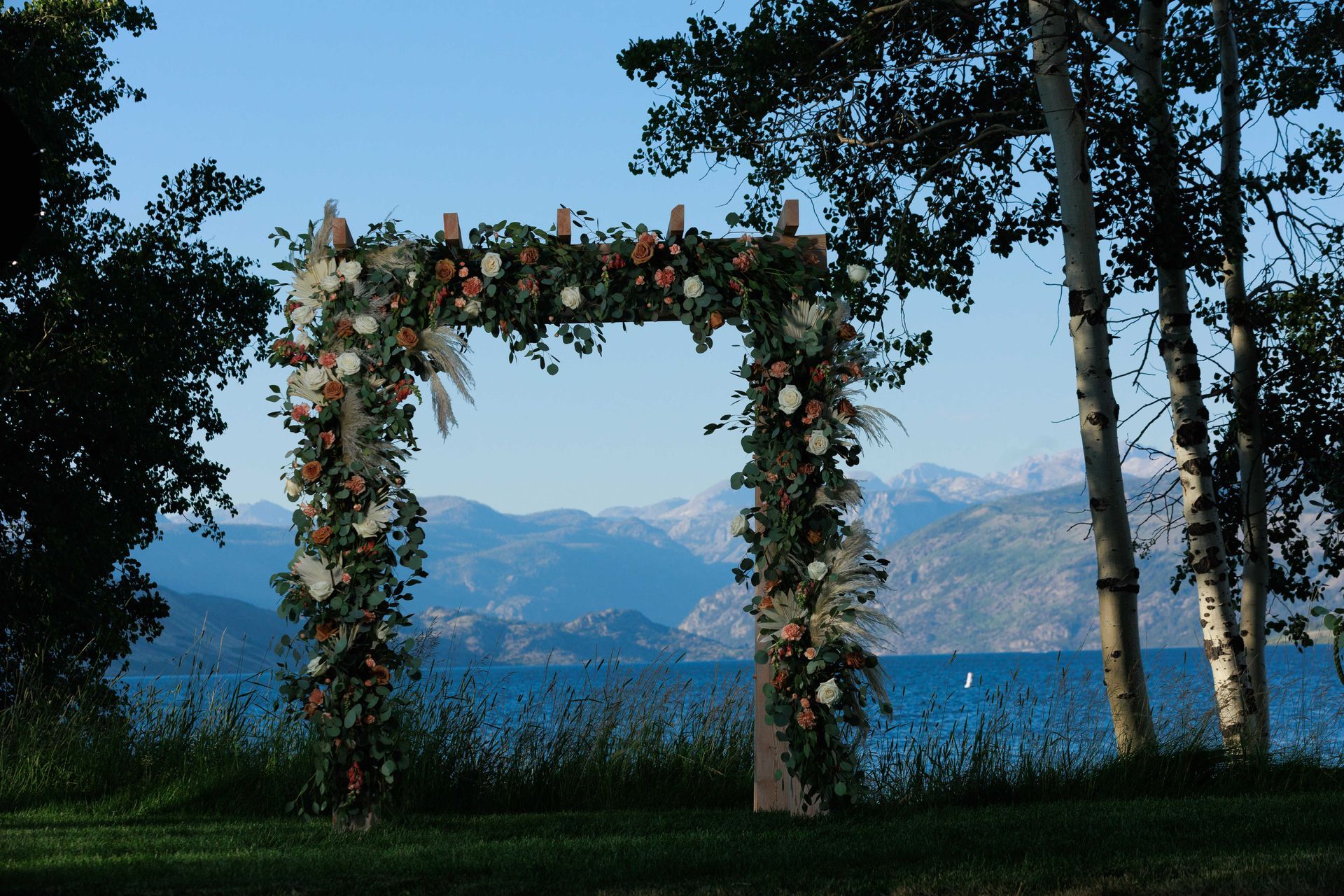 Flower-covered archway by a lake, with mountains and trees in the background.