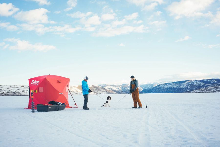 Two people with sled dogs on a snowy lake beside a red tent under a bright blue sky