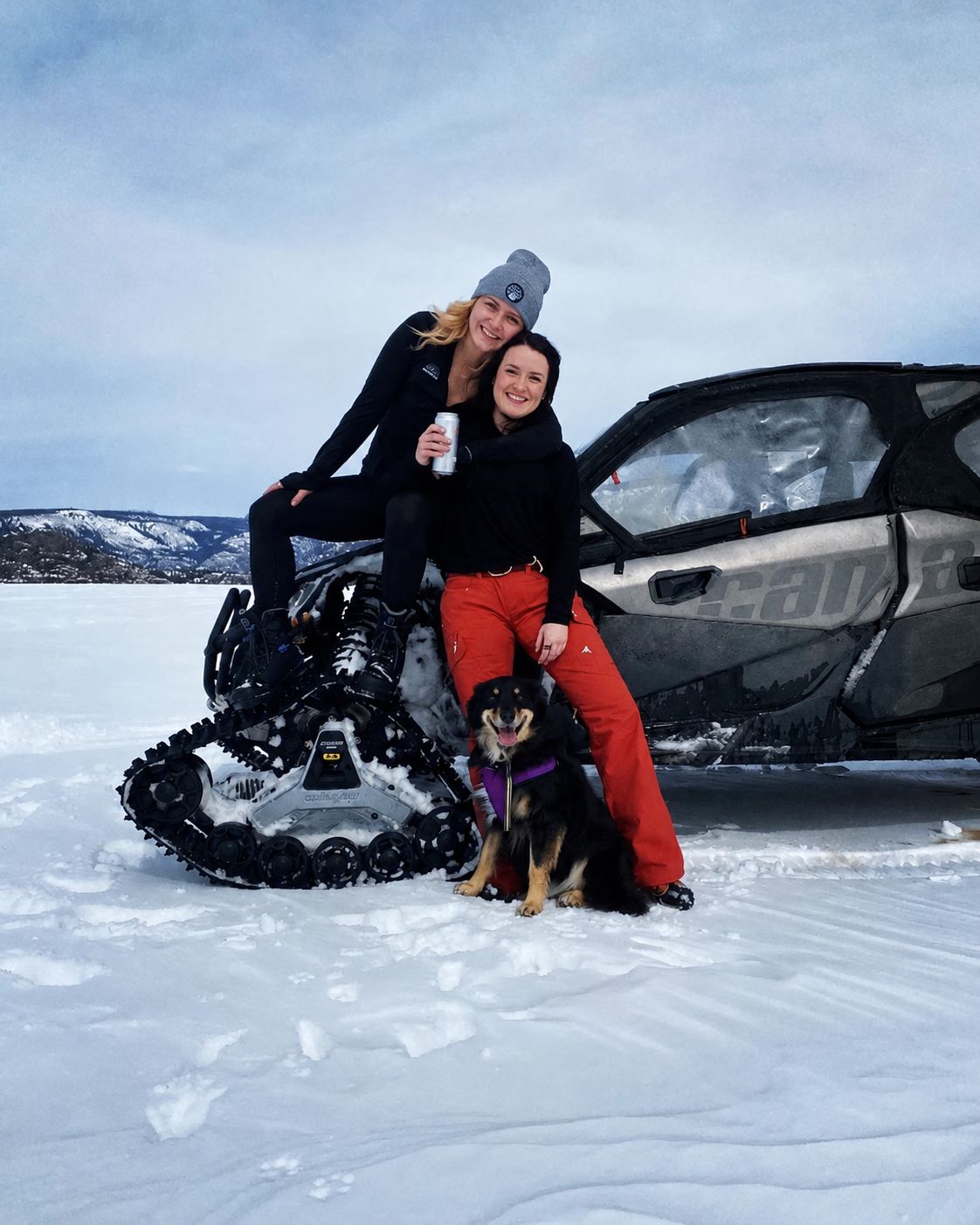 Two people and a dog pose beside a snowmobile on a snowy landscape with mountains in the background