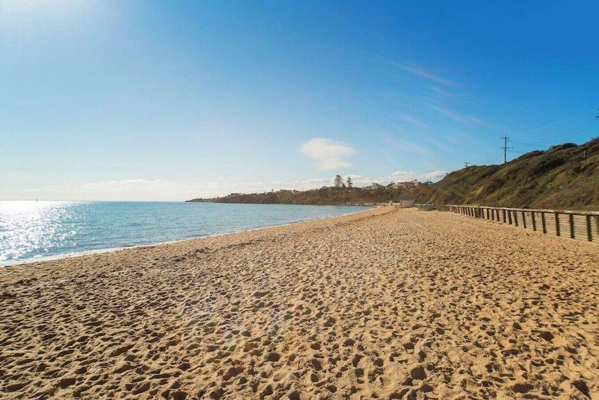 Sandy beach beside calm blue sea, with a sunny coastline and boardwalk along the right side