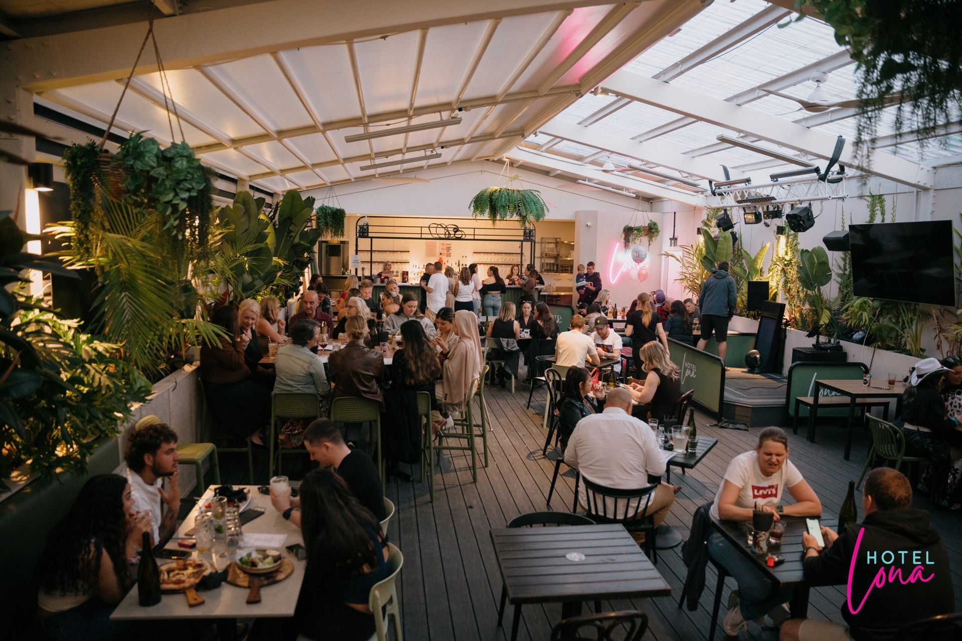 Crowded indoor event with guests dining at tables under string lights in a large tent-like venue