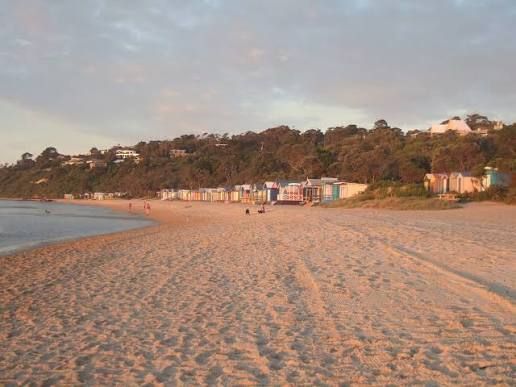 Sandy beach at sunset with calm water, small huts along the shore, and trees on a hillside