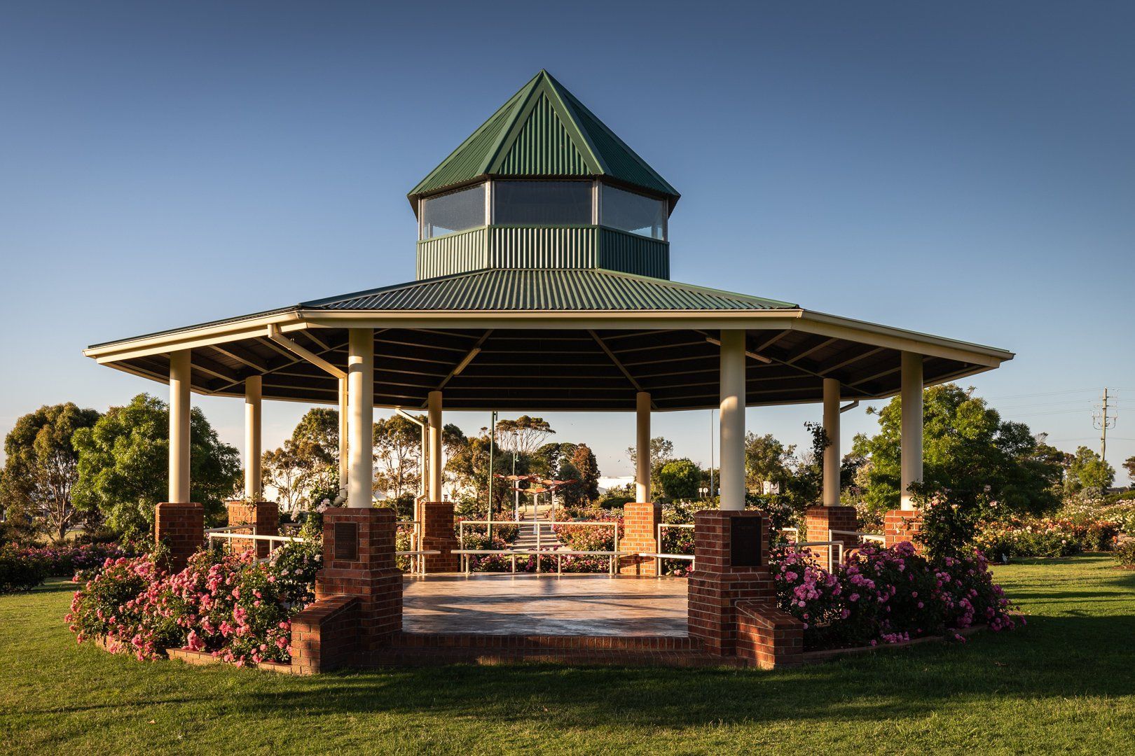 Open-air gazebo with green roof and brick columns in a landscaped park at sunset