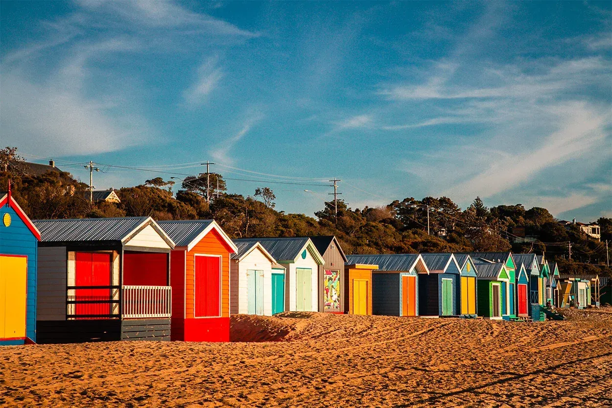 Colorful beach huts lined up on a pebbled shore under a blue sky.