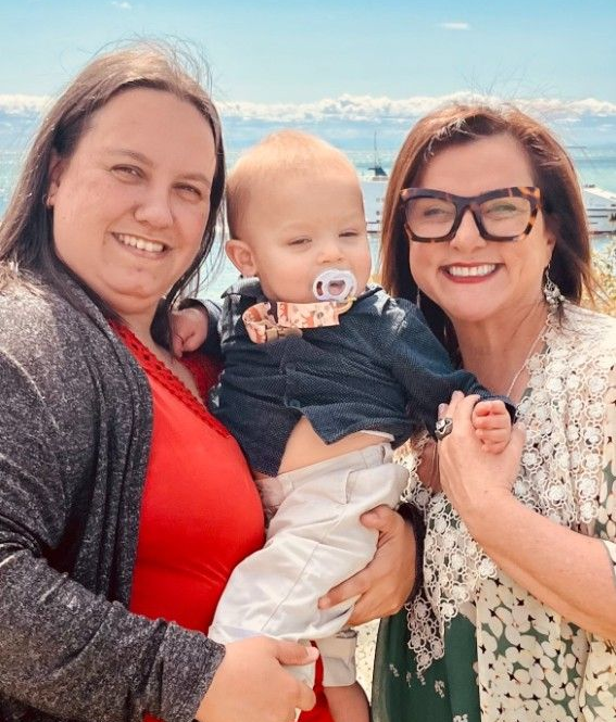 Three people smiling outdoors under a blue sky, with two holding a baby between them.