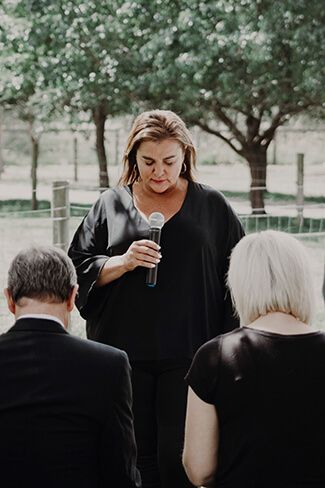 Woman in black dress speaking into a microphone outdoors, facing two seated attendees.