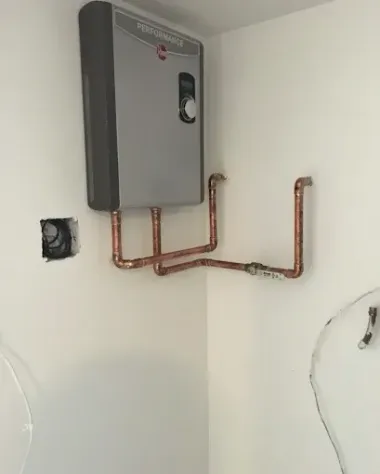 Wall-mounted gray tankless water heater with exposed copper pipes in a white room