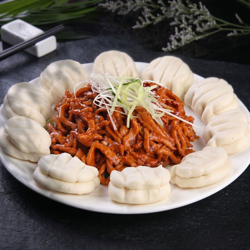 Spicy stir-fried noodles on a white plate, surrounded by steamed buns, topped with shredded scallions