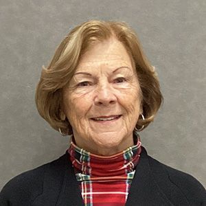 Woman with short, light brown hair smiles, wearing a plaid turtleneck and black blazer against a gray background.