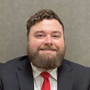 Man in suit, red tie, smiling; background is a neutral wall.